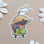 A die-cut sticker featuring a cute cartoon creature with a leafy yellow hat, green coat, and orange scarf