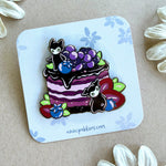 An enamel pin featuring a mixed berry cake with two little spirits.