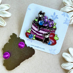 An enamel pin featuring a mixed berry cake with two little spirits showing the front and backside of the pin.